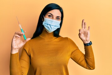 Young caucasian woman wearing medical mask holding syringe doing ok sign with fingers, smiling friendly gesturing excellent symbol