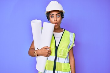 Young african american woman with braids wearing safety helmet holding blueprints scared and amazed with open mouth for surprise, disbelief face