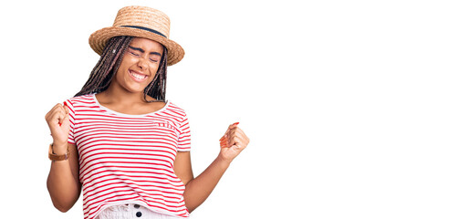 Obraz na płótnie Canvas Young african american woman with braids wearing summer hat very happy and excited doing winner gesture with arms raised, smiling and screaming for success. celebration concept.