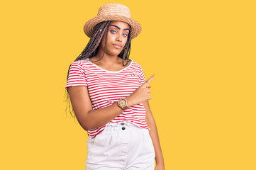 Young african american woman with braids wearing summer hat pointing with hand finger to the side showing advertisement, serious and calm face