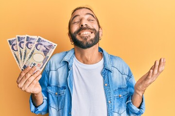 Attractive man with long hair and beard holding 5000 japanese yen banknotes celebrating achievement with happy smile and winner expression with raised hand