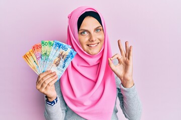 Young caucasian woman wearing islamic hijab holding swiss franc banknotes doing ok sign with fingers, smiling friendly gesturing excellent symbol