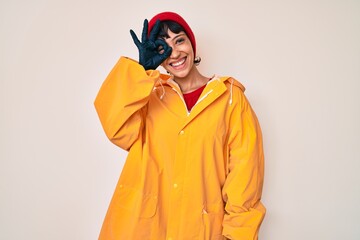 Beautiful brunettte woman wearing yellow raincoat smiling happy doing ok sign with hand on eye looking through fingers