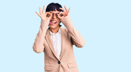 Young brunette woman with short hair wearing business jacket and glasses doing ok gesture like binoculars sticking tongue out, eyes looking through fingers. crazy expression.
