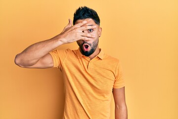 Young man with beard wearing casual clothes peeking in shock covering face and eyes with hand, looking through fingers afraid