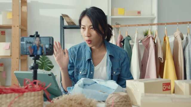 Young Asia lady fashion designer using mobile phone receiving purchase order and showing clothes recording video live streaming online at shop. Small business owner, online market delivery concept.