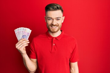 Young redhead man holding 20 polish zloty banknotes looking positive and happy standing and smiling with a confident smile showing teeth