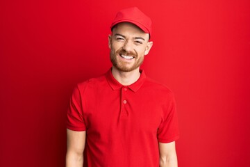 Young redhead man wearing delivery uniform and cap looking positive and happy standing and smiling with a confident smile showing teeth