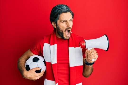 Young hispanic man football hooligan holding ball using megaphone angry and mad screaming frustrated and furious, shouting with anger. rage and aggressive concept.