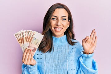 Young brunette woman holding 5000 south korean won banknotes doing ok sign with fingers, smiling friendly gesturing excellent symbol