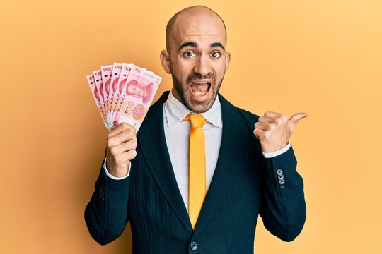 Young hispanic business man wearing business suit holding 100 yuan banknotes pointing thumb up to the side smiling happy with open mouth