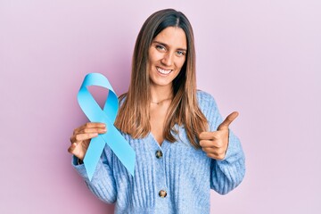 Brunette young woman holding blue ribbon smiling happy and positive, thumb up doing excellent and approval sign