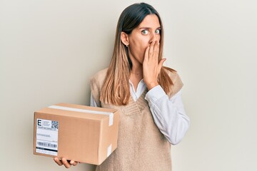 Brunette young woman holding delivery package covering mouth with hand, shocked and afraid for...