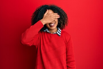 Beautiful african american woman with afro hair wearing sweater and glasses smiling and laughing with hand on face covering eyes for surprise. blind concept.
