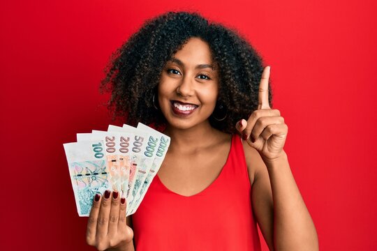 Beautiful african american woman with afro hair holding czech koruna banknotes smiling with an idea or question pointing finger with happy face, number one