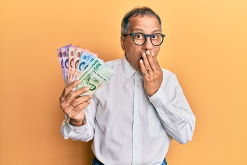 Middle age indian man holding thai baht banknotes covering mouth with hand, shocked and afraid for mistake. surprised expression