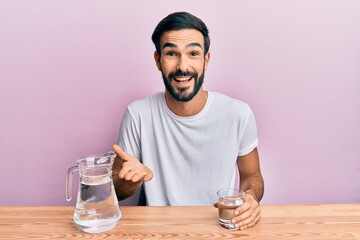 Young hispanic man drinking glass of water sitting on the table celebrating achievement with happy smile and winner expression with raised hand