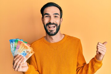 Young hispanic man holding swiss franc banknotes pointing thumb up to the side smiling happy with open mouth