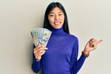 Young chinese woman holding dollars smiling happy pointing with hand and finger to the side