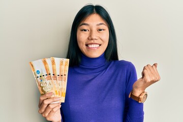 Young chinese woman holding 500 philippine peso banknotes screaming proud, celebrating victory and success very excited with raised arm