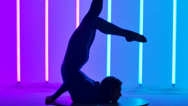 Gymnast in the studio against the background of pink and blue neon lights. The girl does a chest stand, stretches her legs up, sit on the twine. Silhouette. Slow motion. Close up.