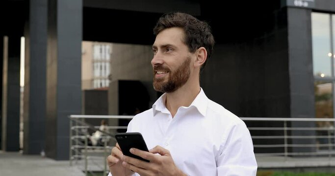 Confident bearded man browsing internet on modern smartphone while standing near office center. Happy man taking break from work on fresh air.
