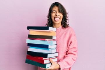 Young hispanic woman holding a pile of books sticking tongue out happy with funny expression.