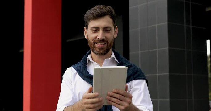 Handsome bearded man in blue sweater sitting outdoors and using portable laptop. Smiling male working on wireless computer on fresh air.
