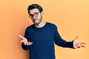 Young hispanic man wearing casual clothes and glasses clueless and confused expression with arms and hands raised. doubt concept.