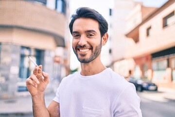 Young hispanic man smiling happy holding cigarette walking at the city.