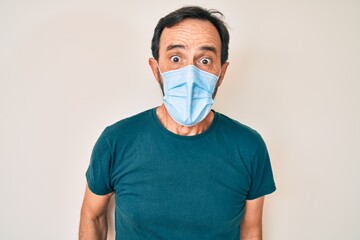Middle age hispanic man wearing medical mask scared and amazed with open mouth for surprise,...