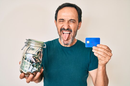 Middle age hispanic man holding credit car and jar with dollars sticking tongue out happy with funny expression.
