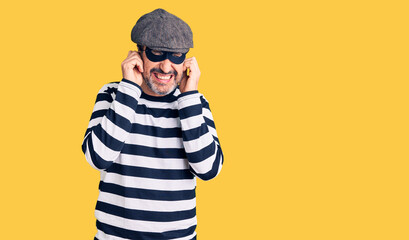 Middle age handsome man wearing burglar mask covering ears with fingers with annoyed expression for...