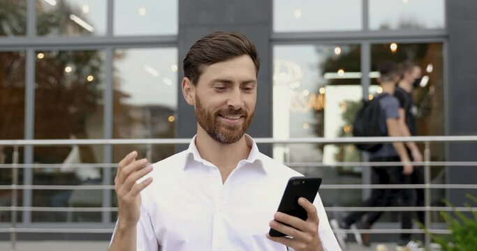 Portrait of successful business man in white shirt suing modern smartphone for video conference out of the office. Bearded male talking and gesturing during online conversation with partners.