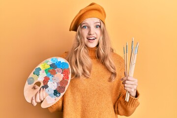 Fototapeta premium Beautiful young caucasian girl holding paintbrush and painter palette celebrating crazy and amazed for success with open eyes screaming excited.