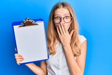 Beautiful young caucasian girl holding clipboard with blank space covering mouth with hand, shocked...
