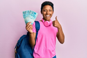 Young african american girl wearing student backpack and holding brazilian real banknotes smiling happy and positive, thumb up doing excellent and approval sign