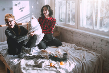 Two young girls fight pillows on the bed in a white room next to the window. Friends or sisters relationships at home