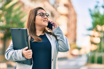 Young plus size businesswoman talking on the smartphone holding binder at the city.