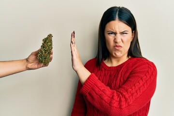 Beautiful brunette woman saying no to marijuana herb skeptic and nervous, frowning upset because of...