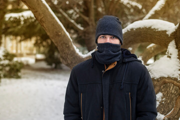 Fototapeta na wymiar The teenager in warm clothes and in a warm hat and scarf covered half of his face because of severe frosts and snow around, the guy is sad because he loves the heat, summer