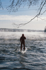 Surfer in a red wetsuit on a board with a paddle floats against the waves. Winter sup surfing on the Vuoksa river. Losev rapids