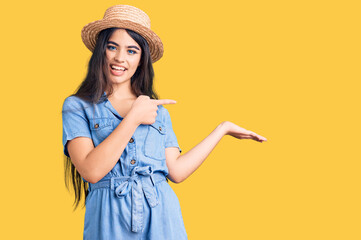 Brunette teenager girl wearing summer hat amazed and smiling to the camera while presenting with hand and pointing with finger.