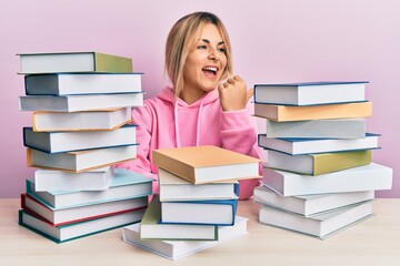 Young caucasian woman sitting on the table with books smiling with happy face looking and pointing to the side with thumb up.