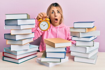 Young caucasian woman holding alarm clock sitting on the table with books afraid and shocked with surprise and amazed expression, fear and excited face.