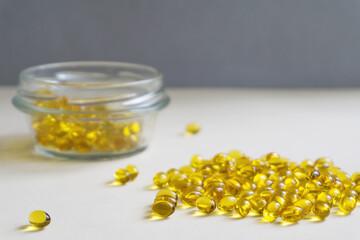 Yellow capsules are scattered on a light background. In the background is a glass jar. Soft focus in the foreground.