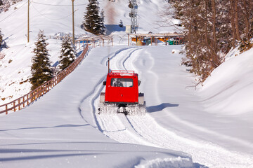 Red snow groomer is going towards the ski resort. Smoking snow vehicle cleared the snow on the road already and left a clean road behind.