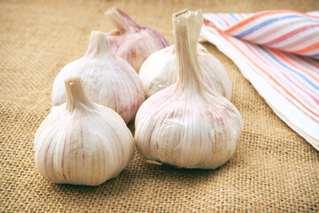 The garlic bulbs lie on a coarse burlap cloth. A small colored-striped towel is spread on the table. Vegetables for preparing various dishes and sauces.