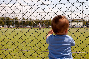Baby boy holding the metal net next to the stadium and watching his brother playing the football game. Little cute baby is learning how to play soccer in his early age.