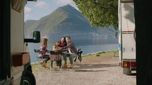 Family eating and sitting on sea shore while traveling by car home outdoors rbbro. Young family with children eats food and sits at table on scenic coast on summer day, camper is nearby. American